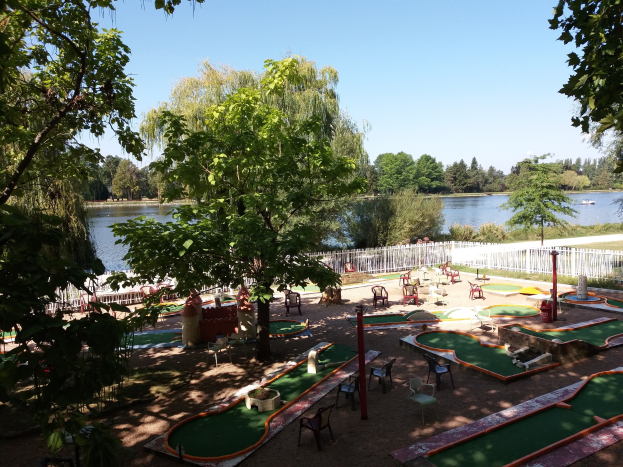 Minigolfplatz in einem Park mit grünem Gras, Bäumen und einem Zaun, umgeben von verstreuten Stühlen und Tischen, mit einem Gewässer und einem Boot im Hintergrund unter dem Himmel.