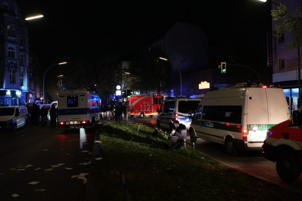 Eine Gruppe von Polizeiwagen steht nachts am Straßenrand geparkt, mit Menschen auf dem Bürgersteig, Gras, Pfählen, Lichtern, Verkehrsampeln, Schildern, Bäumen, Gebäuden mit Fenstern und dem Himmel im Hintergrund.