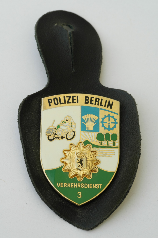 Ein schildförmiges Polizeibadge mit "Polizei Berlin" in schwarzer fettgeschriebener Schrift auf einem blauen Hintergrund mit einem weißen Stern und Rahmen, das auf einer Oberfläche platziert ist.