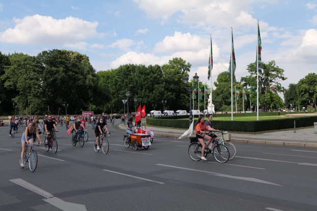 Eine Gruppe von Menschen, die auf Fahrrädern eine von Bäumen gesäumte Straße mit Laternenmasten und Fahnenmasten entlangfahren, während der Pariser Fahrradtag 2016 stattfindet, mit Fahrzeugen und einer bewölkten Kulisse im Hintergrund.