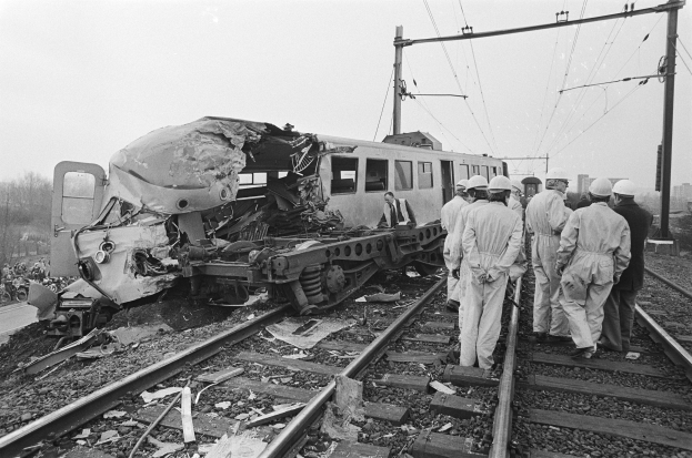 Ein Schwarz-Weißs Bild einer Zugkollision mit Einsatzkräften in Helmen, die um das Wrack herumstehen, vor einer Kulisse aus Strommästen, Bäumen, Gebäuden und einem klaren Himmel.