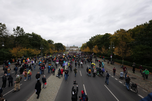 Eine große Gruppe von Menschen, die eine von B√∞umen gesäumte Straße in Berlin entlanggehen, mit Kameras in der Hand, einem Gebäude und einem klaren Himmel im Hintergrund.
