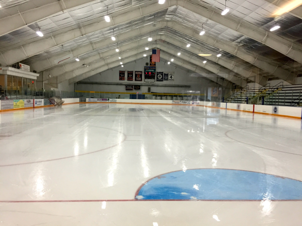 Große Indoor-Eisfläche mit Deckenbeleuchtung, Werbetafeln, Treppen mit Geländern und einigen Menschen in einer Sportanlage.