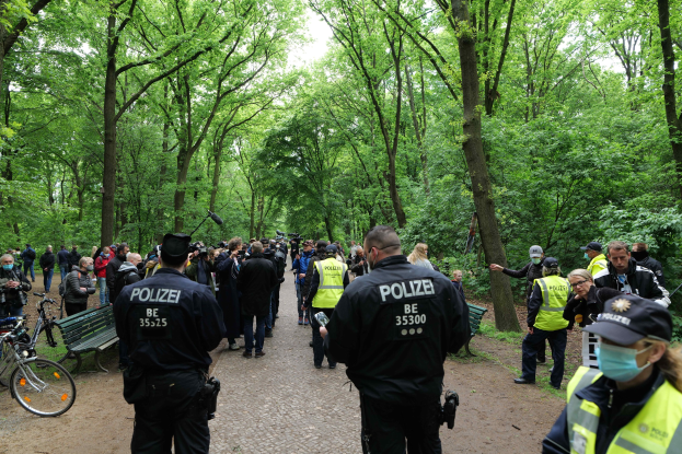 Eine Gruppe von Polizisten steht vor einer Menschenmenge, mit Fahrrädern und einer Bank im Vordergrund und Bäumen im Hintergrund.