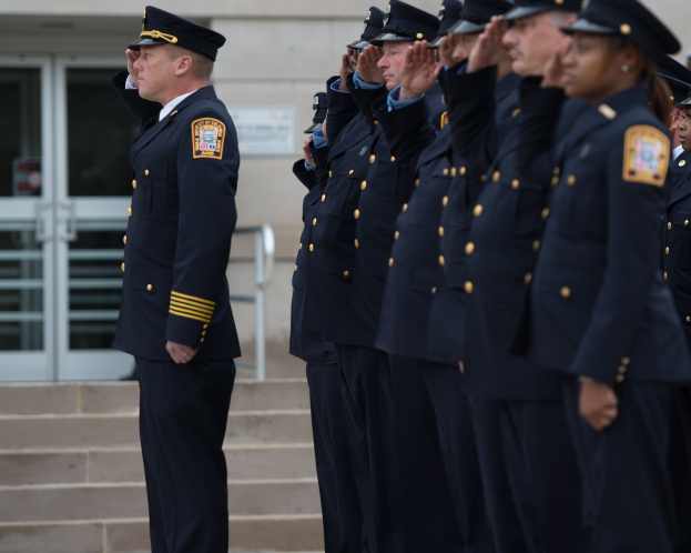 Gruppe von Polizisten in Uniform salutiert im Glied vor einem Gebäude mit Glastüren und Treppe.