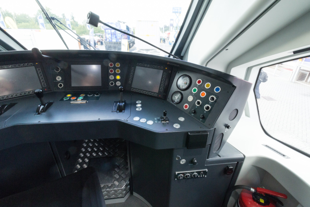 Cockpit eines Zuges mit Feuerlöscher im Vordergrund, das eine Glasfensteransicht von Menschen, Fahrzeugen, Bäumen, Masten und anderen Gegenständen außerhalb zeigt.