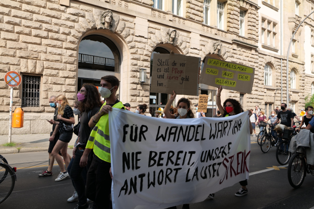 Eine Gruppe von Menschen marschiert bei einer Demonstration gegen die Anti-Kriegsbewegung in Berlin, einige halten Schilder und andere fahren Fahrräder, im Hintergrund ein Gebäude mit Bögen, Säulen und Skulpturen.