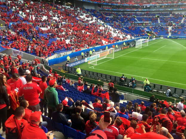 Eine große Menschenmenge in einem Stadion schaut einem Fußballspiel zu, mit Sitzplätzen und Stehplätzen, Bannern, einem Torpfosten und Deckenlampen.