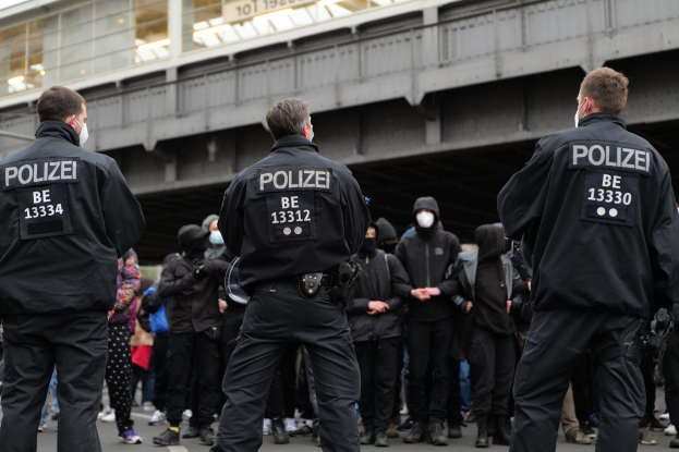 Eine Gruppe von Polizisten in Uniform steht vor einer Menge von Menschen in schwarzen Uniformen und Masken, mit einer Brücke und einem Gebäude im Hintergrund, was eine städtische Umgebung während einer Demonstration vermuten lässt.