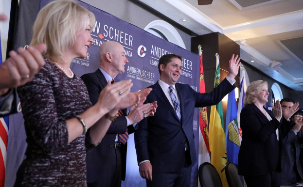 Eine Gruppe von Menschen, die vor Flaggen stehen und klatschen, mit einem Tisch mit Gegenständen rechts, Stühlen, einer Fahne und einer Wand im Hintergrund und Lichtern und einem Ventilator auf dem Dach.