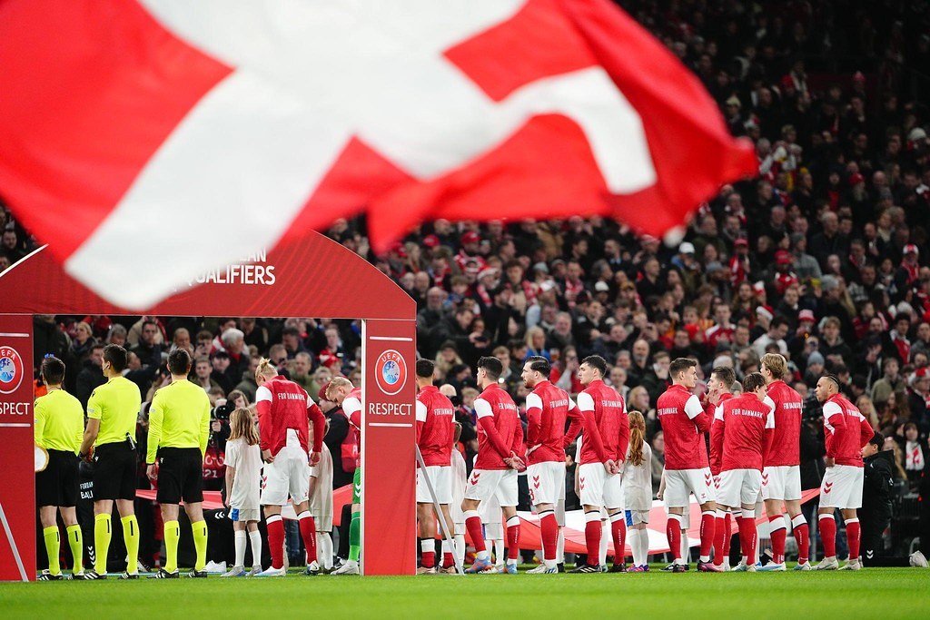 Eine Gruppe von Menschen steht auf einem Fußballfeld mit einer roten und weißen Flagge im Vordergrund, einem Bogen mit der Aufschrift "Bayern Munich vs Bayern Munich Betting Tips & Preview" im Hintergrund und einer großen Menge im Stadion.