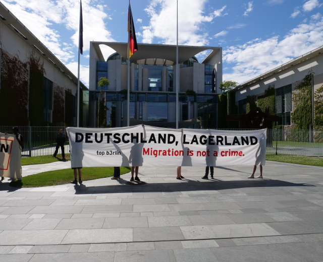 Eine Gruppe von Menschen hält ein Transparent mit der Aufschrift "Deutschland, Lagerland Migration ist kein Verbrechen" vor einem eingezäunten Gebäude mit Bäumen und Fahnen unter einem bewölkten Himmel.