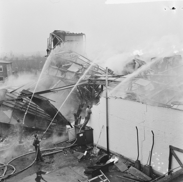 Schwarzes und weißes Foto von Feuerwehrleuten auf einem Gebäudedach bei der Brandbekämpfung, während Menschen unten Rohre und eine Leiter halten.