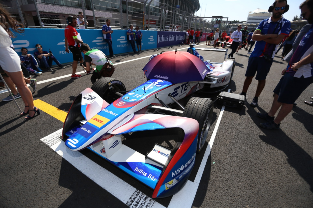 Formula E Auto mit weißer, blauer, roter und schwarzer Lackierung auf einer Rennstrecke geparkt, umgeben von Menschen, mit einem Schirm im Vordergrund und Gebäuden, Bäumen und einem klaren blauen Himmel im Hintergrund.