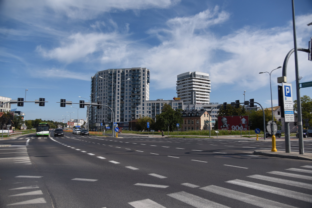 Stadtstraße mit fahrenden Fahrzeugen, Laternen, Verkehrsampeln, Schildern, Bäumen, Gebäuden mit Fenstern und einem bewölkten Himmel.