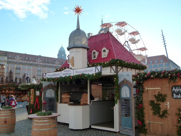 Ein kleines rotes Dachgebäude mit einem Riesenrad davor, umgeben von Menschen und Festobjekten, vor Gebäuden und Bäumen unter einem bewölkten Himmel bei einem Oktoberfest.
