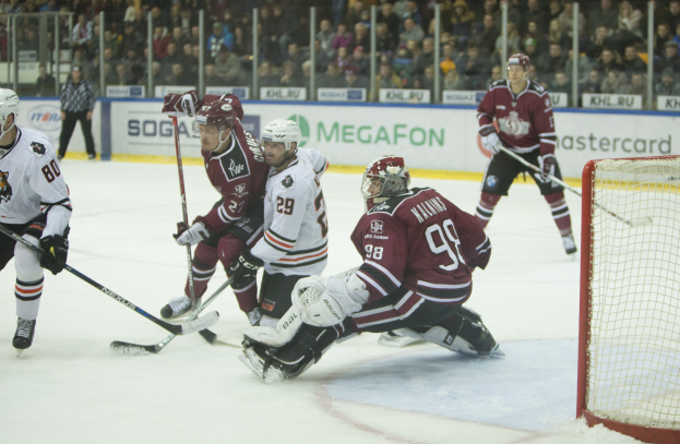 Gruppe von Menschen, die Hockey auf einem Eisstadion spielen, mit Torpfosten auf der rechten Seite, tragen Helme und halten Stöcke, Zuschauer auf Tribüne mit Bannern im Hintergrund.