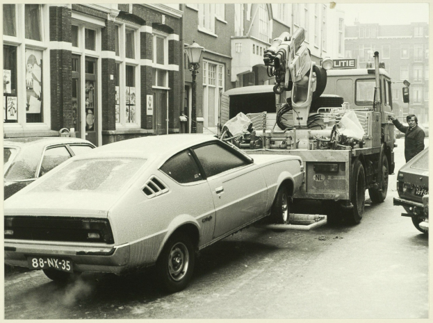 Ein Schwarz-Weiß-Foto eines Autos, das von einem Abschleppwagen auf einer Straße mit anderen Fahrzeugen, einer Person in der Nähe und Gebäuden mit Fenstern im Hintergrund abgeschleppt wird.