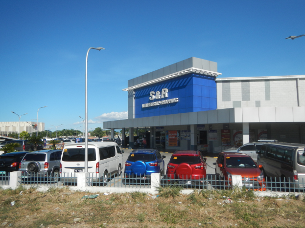 Ein Autohaus mit vorne geparkten Fahrzeugen, umgeben von einem Metallzaun, Gras, Pflanzen, Bäumen, Straßenlaternen und einem bewölkten Himmel im Hintergrund.