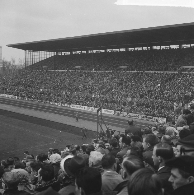 Schwarzes und weißes Foto einer vollen Tribüne mit Zuschauern, die ein Fußballspiel verfolgen, mit Bannern, Pfählen, einem Schuppen, Bäumen, einem Turm und einem bewölkten Himmel.
