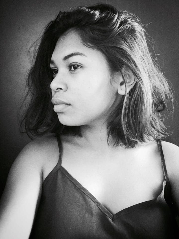 Eine selbstbewusste Frau in einem schwarzen Kleid posiert in einem Schwarz-Weiß-Foto, ihr glatter Bob und das natürliche Make-up unterstreichen ihre selbstbewusste Haltung gegen eine Wand.