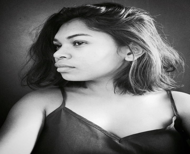 Eine selbstbewusste Frau in einem schwarzen Kleid posiert in einem Schwarz-Weiß-Foto, ihr glatter Bob und das natürliche Make-up unterstreichen ihre selbstbewusste Haltung gegen eine Wand.