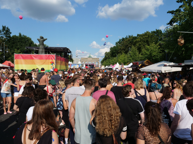 Eine große Menschenmenge geht eine Straße mit Zelten, Bäumen, Pfählen, Lichtern und einer Statue entlang, mit Gebäuden und einem Himmel voller Wolken und Ballons im Hintergrund beim Christopher Street Day Festival in Berlin.