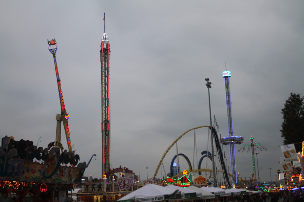 Eine nächtliche Faschingszene mit einer Menge Menschen um beleuchtete Fahrgeschäfte, Pfähle und Bäume unter einem bewölkten Himmel.