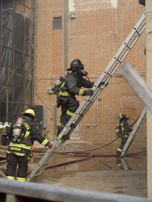Feuerwehrleute in Helmen und Sauerstoffflaschen klettern an einer Leiter vor einem Backsteingebäude mit Rohren auf dem Boden und einer Metallstange am unteren Ende, mit einem anderen Gebäude mit Fenstern und einem Netz im Hintergrund.