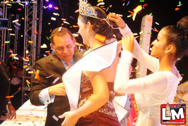 Ein Mädchen auf der Bühne mit einer Krone, mit einem anderen Mädchen, das sie von hinten anpasst, während ein Mann in einem schwarzen Anzug ihr die Hand schüttelt, vor einem dunklen Hintergrund mit Metallrahmen.