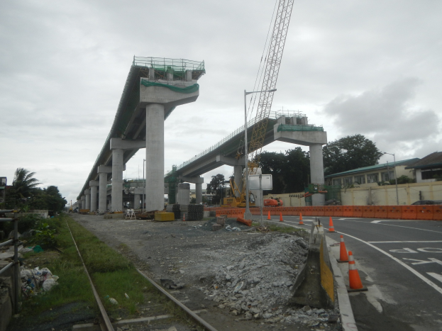 Baustelle mit einer Brücke im Hintergrund, Straße mit Absperrbaken markiert, verstreute Steine und Gras, Bahnschiene auf der linken Seite, Bäume und Gebäude säumen die Straße und ein bewölkter Himmel.