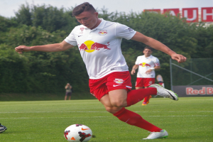 Ein Mann in einem Fußballtrikot tritt einen Ball auf einem grünen Feld mit Bäumen und einem klaren blauen Himmel im Hintergrund, mit einem Schild, auf dem "RB Leipzig" steht.