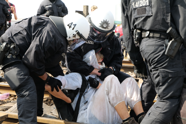 Polizisten in Einsatzausrüstung verhaften einen Mann in einem weißen Kleid auf einem Bahngleis, mit anderen Beamten in schwarzen Uniformen und Helmen.