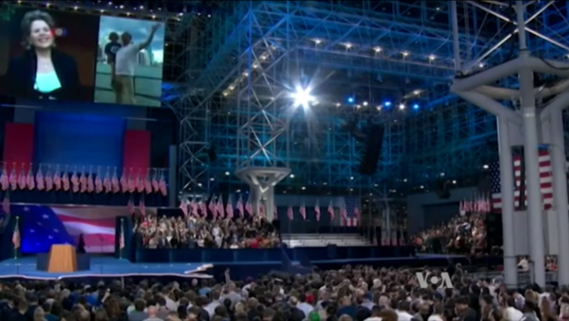 Eine große Menschenmenge sitzt vor einer Bühne mit einem Podium, einem Mikrofon, Flaggen, einem Bildschirm und einer Lichtanlage und wartet auf eine Präsidentschaftsdebatte.