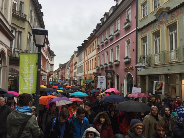 Große Gruppe von Menschen, die auf einer Stadtstraße mit Schirmen gehen, einige tragen Taschen und Schilder, mit Gebäuden auf beiden Seiten und einem Laternenpfahl im Vordergrund.