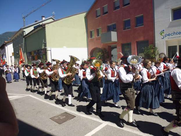 Eine Gruppe von Menschen in traditioneller bayrischer Tracht, die Musikinstrumente spielen, während sie eine Straße mit Gebäuden, einigen Fahnen, einem Hügel und einem blauen Himmel entlanggehen.
