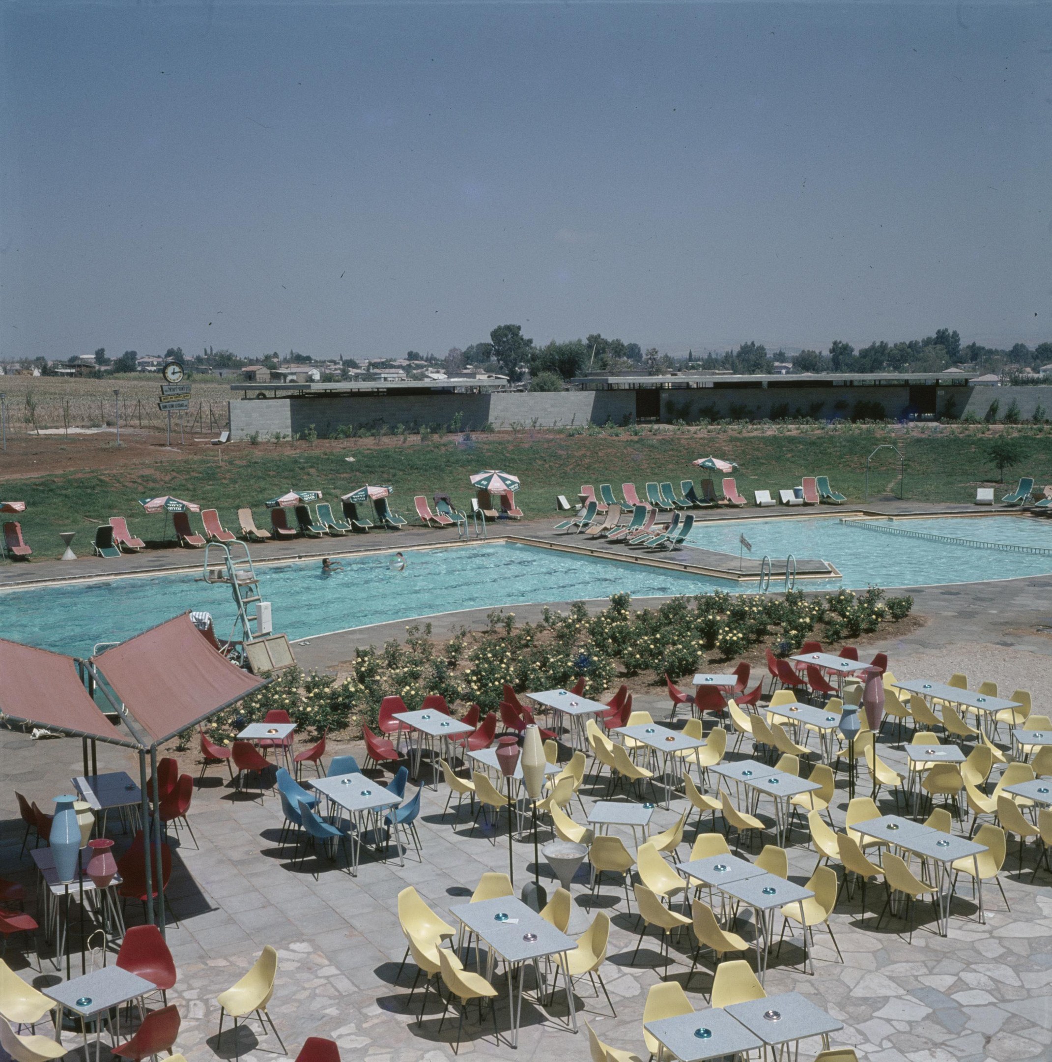 Freibad mit Tischen, Stühlen, Pflanzen, Blumen, Sonnenschirmen, Pfählen, einem Zaun, einer Wand, Bäumen, Gebäuden und einer bewölkten Himmel.