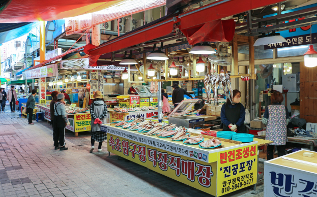Ein belebter Straßenmarkt in Seoul, Südkorea, mit Menschen, die gehen, Ständen, die Artikel wie Lichter und Schilder ausstellen, und Gebäuden unter einem klaren blauen Himmel.