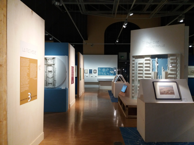 Ein volles Kunstausstellung-Zimmer mit gerahmten Fotos auf Tischen, Texttafeln an Wänden, Deckenlampen und einem Teppichboden im Museum of Modern Art.