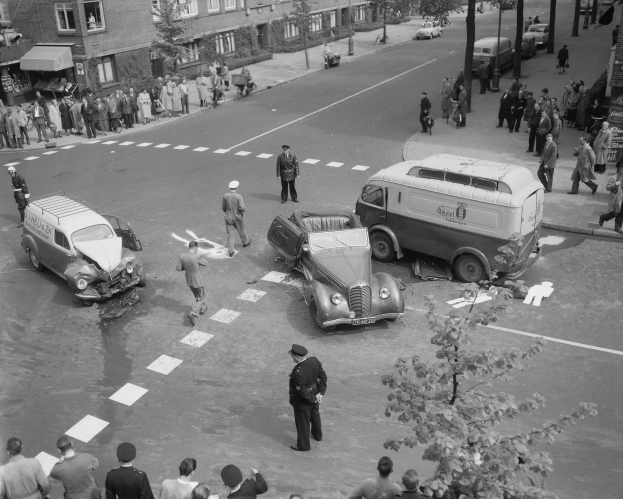 Eine Schwarz-Weiß-Szene eines Autounfalls an einer Straßenkreuzung mit mehreren Fahrzeugen, darunter ein Van, einer Gruppe von Menschen drumherum und im Hintergrund Gebäude mit Fenstern, Bäumen, Pfählen und Texttafeln.