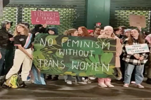 Eine Gruppe von Menschen hält ein Transparent mit der Aufschrift "Kein Feminismus ohne Trans-Frauen" und verschiedene Schilder in einem öffentlichen Raum.