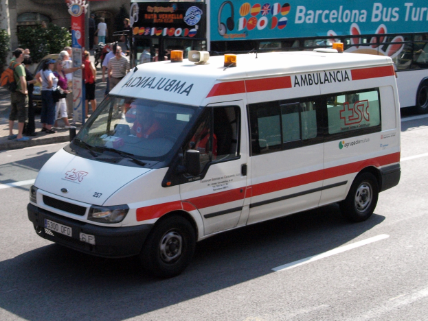 Rettungswagen fährt auf einer Straße neben einem Bus, mit Menschen, die auf dem Gehweg neben dem Bus stehen, Gebäude und Bäume im Hintergrund bei klarem blauem Himmel.