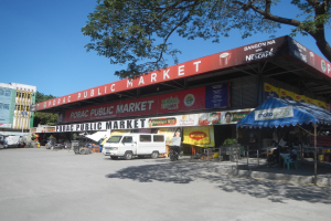 Öffentlicher Markt mit einem vor ihm geparkten Van, umgeben von Gebäuden, Geschäften, Schildern, Fahrzeugen auf der Straße, Personen unter einem Zelt sitzend, Geländern, Bäumen und einem klaren blauen Himmel.