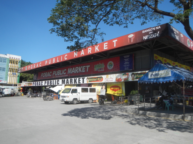 Öffentlicher Markt mit einem vor ihm geparkten Van, umgeben von Gebäuden, Geschäften, Schildern, Fahrzeugen auf der Straße, Personen unter einem Zelt sitzend, Geländern, Bäumen und einem klaren blauen Himmel.