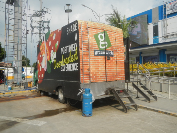 Lkw vor einem Gebäude mit einer Tafel, auf der "Teilen Sie ein positiv überladenes Erlebnis" steht, mit Stufen, einem Zylinder auf dem Boden, Geländern, Strom- und Lichtmasten, Bäumen und einem bewölkten Himmel.
