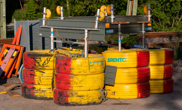 Ein Haufen gelber und roter Reifen auf einem Schotterfeld umgeben von verstreuten Metallobjekten, Brettern und anderem Schutt, mit Bäumen, Pflanzen und einem Schuppen im Hintergrund.