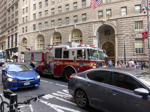 Ein rotes Feuerwehrauto auf einer Straße in New York City mit fahrenden Fahrzeugen, Fußgängern auf dem Gehweg, parkenden Fahrrädern und Gebäuden mit Fenstern, Laternenmasten und Fahnenmasten im Hintergrund.