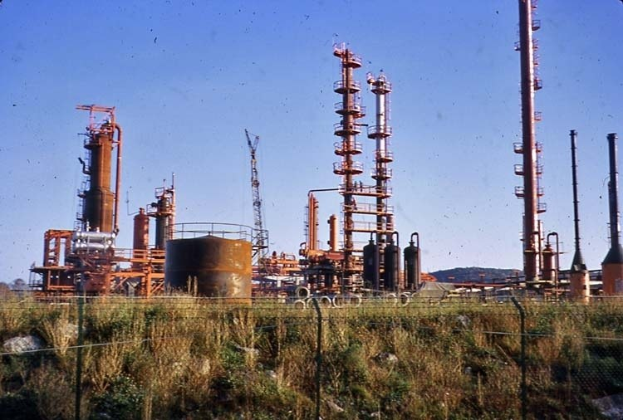 Ölraffinerie mit Tanks und Equipment in einer Grasfläche, umgeben von einem Zaun, mit Hügeln und einem klaren blauen Himmel im Hintergrund.