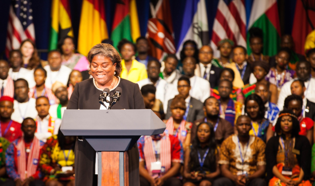Frau spricht an einem Rednerpult mit Mikrofonen vor einem aufmerksamen Publikum, mit sichtbaren Flaggen im Hintergrund.