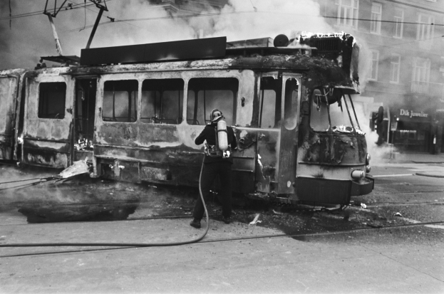 Schwarz-weißes Bild eines Mannes in einem Helm, der eine Pfeife h├Ąlt und neben einer ausgebrannten Stra├čenbahn auf der Stra├če steht, mit Rauch aufsteigend, ├ťberkopfdr├Ąhten und einem Gebäude im Hintergrund.